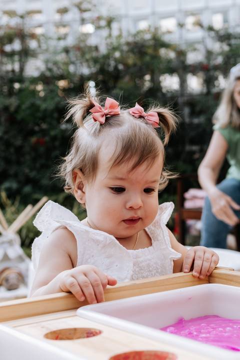 Olívia brincando durante sua festa de 1 ano, usando vestido branco e laços rosas no cabelo.'