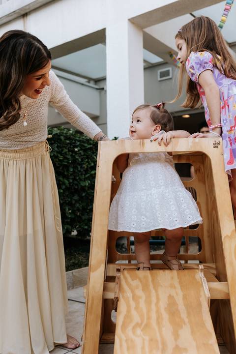 Olívia sentada brincando durante sua festa de 1 ano, enquanto a mãe acompanha ao lado.'