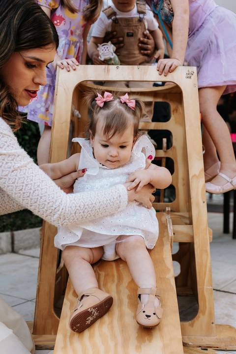 Olívia sentada em uma cadeirinha de madeira enquanto recebe a ajuda de um adulto durante a festa.'