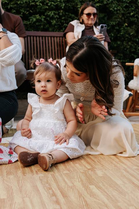 Olívia sentada no colo da mãe durante a festa, observando os convidados.'