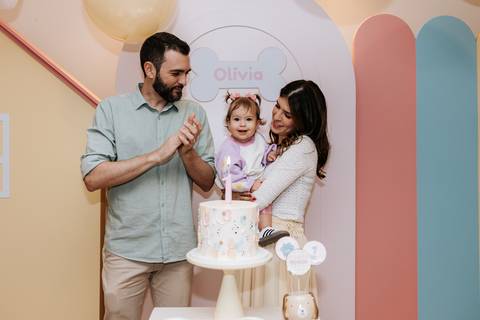 Olívia comemorando seu aniversário ao lado dos pais enquanto se aproxima do bolo.'