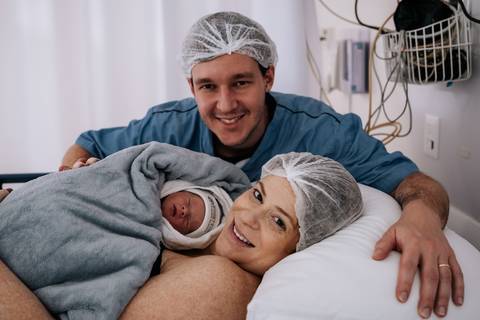 Fabricio abraça Jessica enquanto os dois seguram a recém-nascida Maria Clara, celebrando juntos os primeiros instantes em família.'
