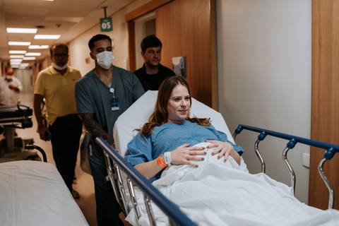 Jessica é conduzida em uma maca pelo corredor da maternidade, acompanhada por Fabricio, familiares e profissionais da equipe médica.'
