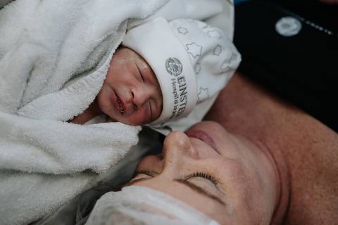Jessica contempla Maria Clara nos primeiros minutos de vida, com a bebê aconchegada em uma manta e usando a touca da maternidade.'