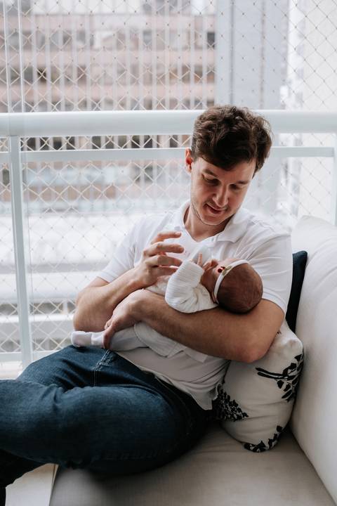 Fabrício segura Maria Clara no colo e observa a filha com ternura enquanto ela descansa confortavelmente em seus braços.'