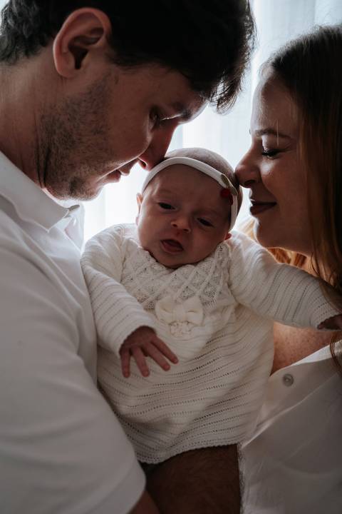 Maria Clara aparece entre os pais, com Jessica e Fabrício aproximando os rostos para contemplar a recém-nascida em um momento de conexão familiar.'