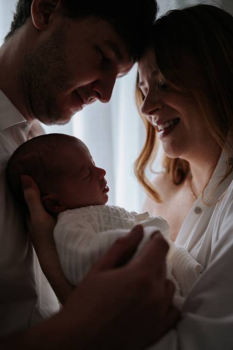 Retrato em contra-luz do casal Jessica e Fabricio admirando a recém-nascida Maria Clara no colo.'