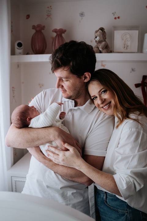 Retrato aconchegante da família no quarto com Fabricio segurando Maria Clara ao lado de Jessica.'