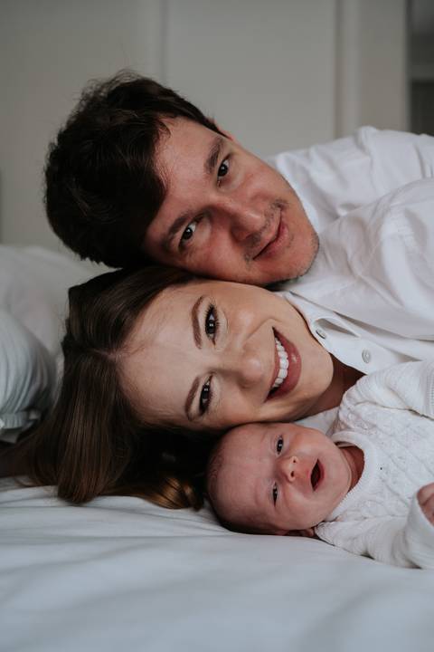 casal sorrindo deitados com a bebê Maria Clara na cama.'