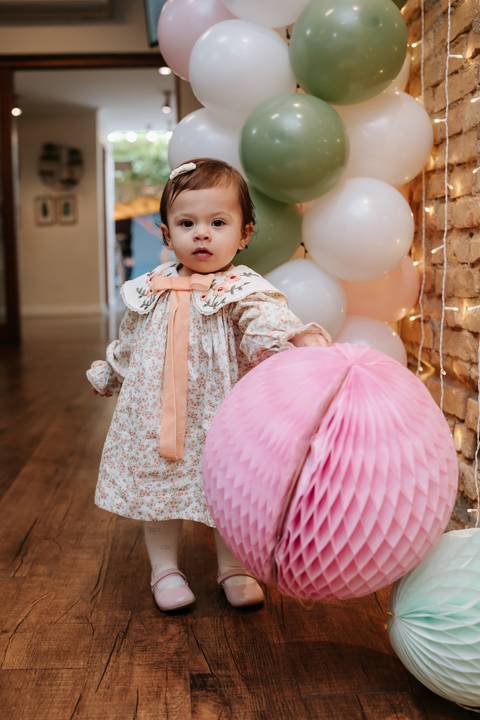 Madalena posa ao lado de uma grande decoração de papel rosa, usando um vestido delicado para celebrar seu primeiro aniversário.'