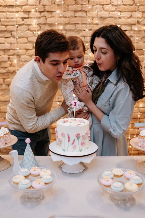 Mariana e Pedro Henrique beijam Madalena durante o parabéns, compartilhando um momento cheio de carinho diante do bolo.'