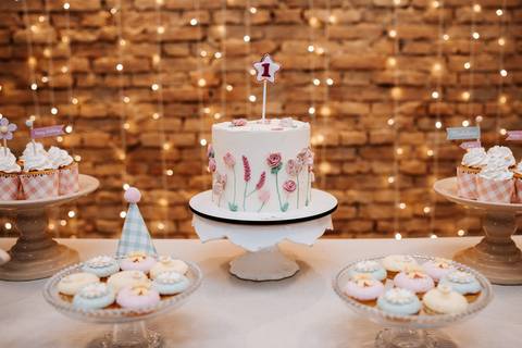 Bolo de aniversário decorado com flores delicadas e uma vela de 1 ano, em meio às luzes aconchegantes da festa.'