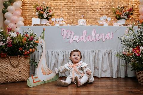 Madalena posa diante da mesa principal decorada para seu primeiro aniversário, com flores, balões e o tema gansinho.'