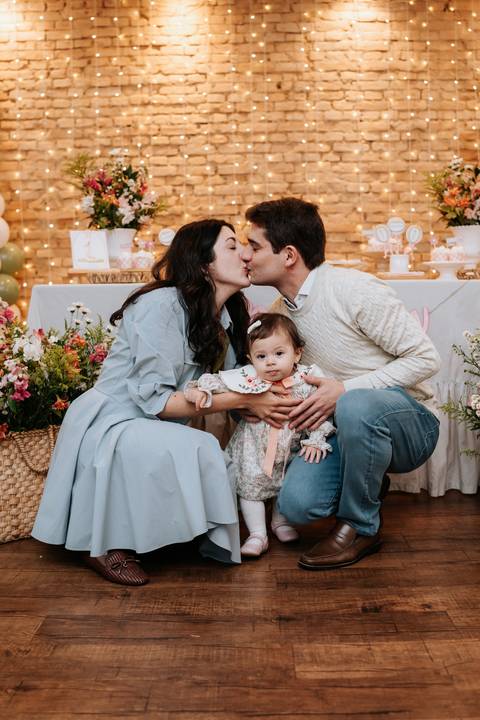 Mariana e Pedro Henrique trocam um beijo carinhoso enquanto seguram Madalena, celebrando juntos esse momento especial.'