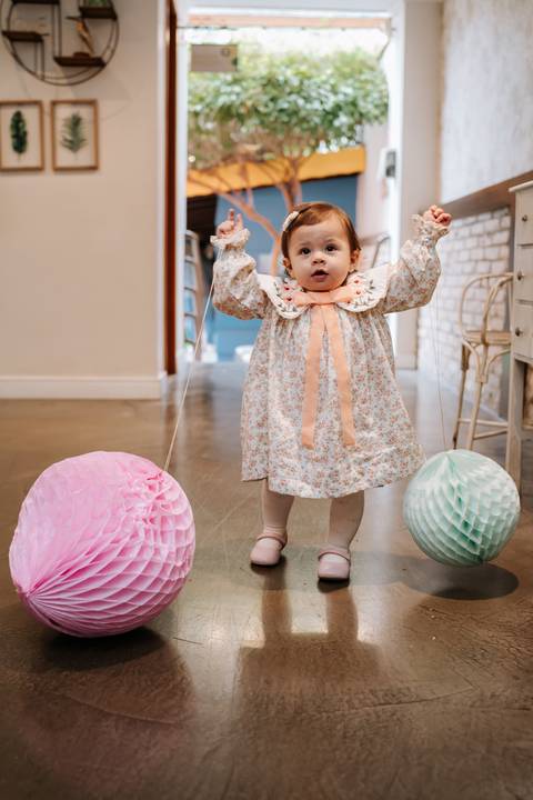Madalena brinca com os braços levantados, usando seu vestido floral em meio à decoração delicada da festa.'