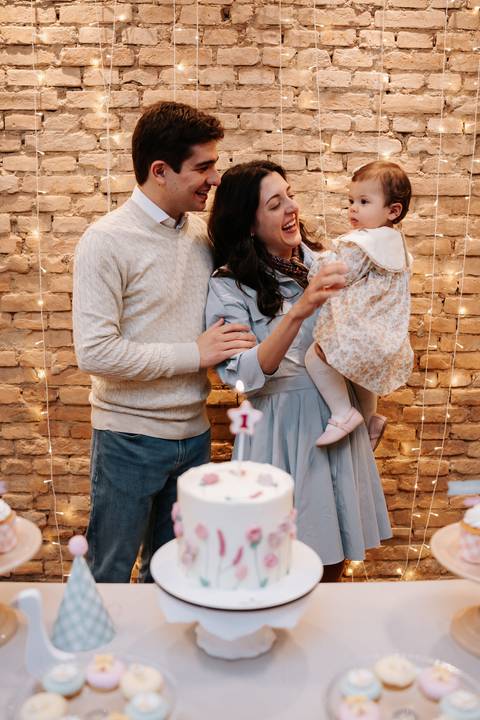 Mariana e Pedro Henrique erguem Madalena diante do bolo para celebrar seu primeiro aniversário em família.'
