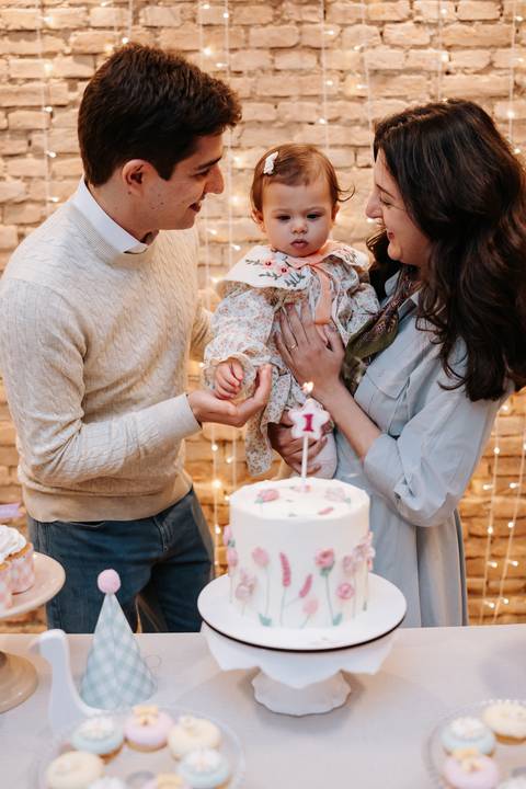 Mariana e Pedro Henrique erguem Madalena diante do bolo para celebrar seu primeiro aniversário em família.'