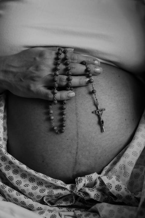 Foto em preto e branco em close na barriga de Marina com o terço repousado sobre ela.'