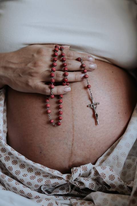 Detalhe das mãos sobre a barriga de Marina segurando um terço.'