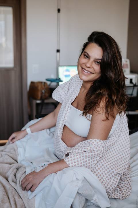 Marina sorridente sentada na cama da maternidade com a barriga da espera de José Joaquim à mostra.'