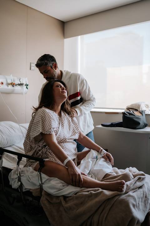 José Carlos em pé ao lado da cama hospitalar apoiando e conversando com Marina.'