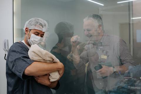 José Carlos junto ao vidro da sala de observação a apresentar o filho aos familiares que tiram fotos.'
