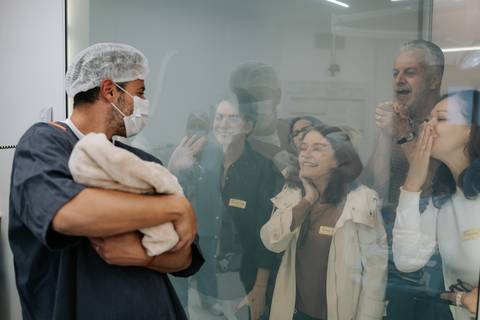 Família a vibrar e a comemorar através do visor de vidro ao conhecer o recém-nascido.'