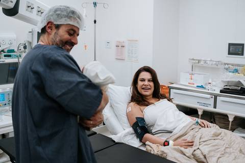 Marina sentada na cama da sala de recuperação a sorrir enquanto José Carlos segura o bebé ao lado.'