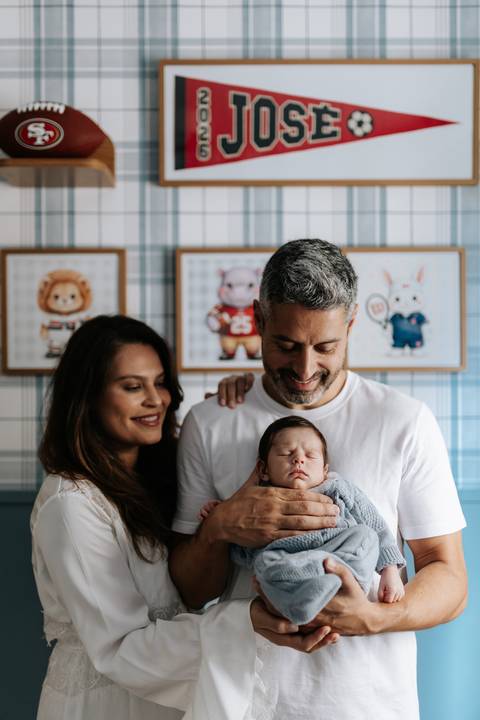 Casal reunido e sorrindo com ternura enquanto seguram José Joaquim adormecido em frente aos quadrinhos.'