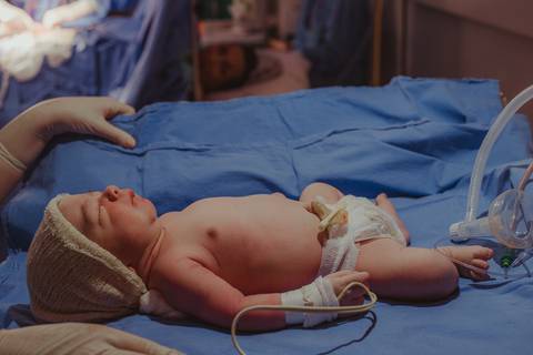 bebê recém-nascido em berço aquecido, em perspectiva à mãe na maca, que observa os primeiros cuidados com o bebê'