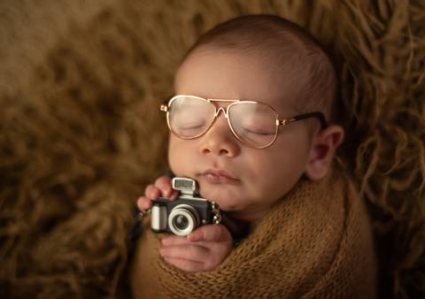 bebê segurando mini câmera e com óculos de grau em ensaio newborn de recém-nascido'