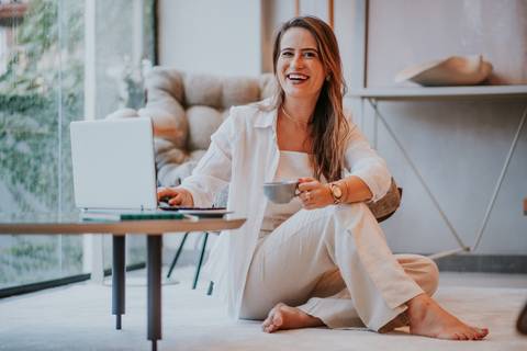 Mulher sentada no chão com notebook e xícara, em momento descontraído e profissional.


'
