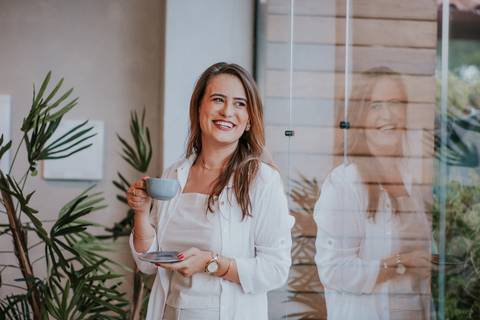 Reflexo de mulher sorridente em vidro, compondo cena estética com plantas ao fundo.


'
