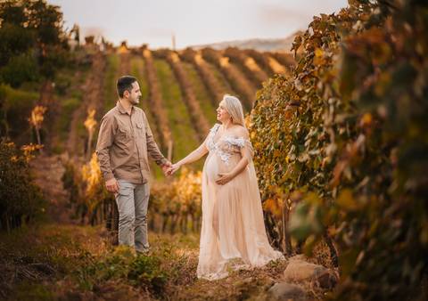 Casal entre os vinhedos trocando carinho e sorrisos, em um retrato de cumplicidade e amor na gestação.'