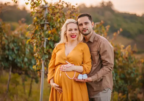 Casal sorrindo, com terço e sapatinho azul sobre a barriga da gestante. Detalhes que expressam fé e espera.'