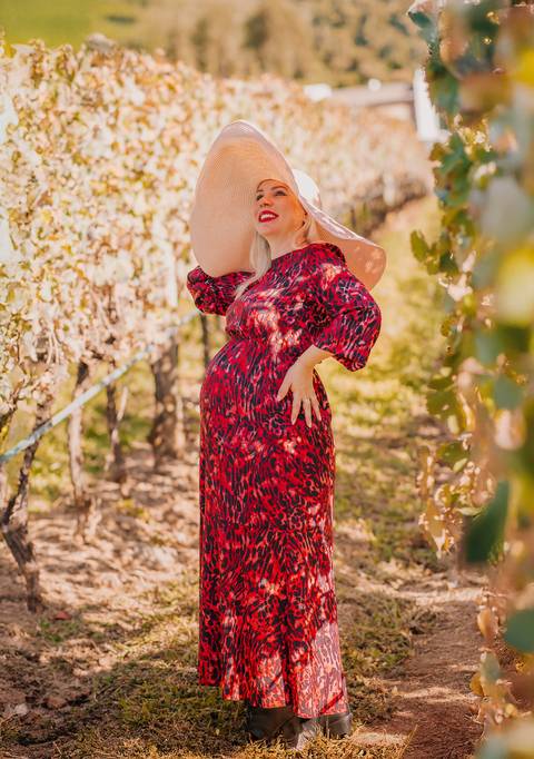Ensaio de gestante com chapéu e expressão de plenitude, cercada por parreirais em tom dourado na serra gaúcha.'