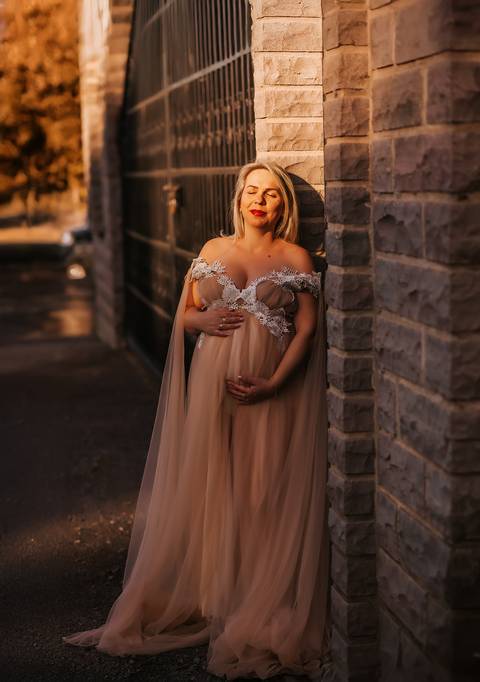 Gestante com vestido esvoaçante bege encostada em parede de pedra, em um momento de contemplação e conexão com o bebê.'