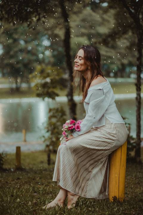 Mulher sorrindo de olhos fechados em meio à chuva e à natureza, ensaio pessoal feminino em dia chuvoso.'