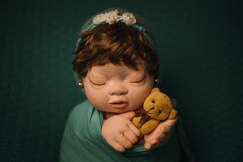 Boneca reborn embrulhada em tecido verde e mãos sob o queixo, utilizada para aulas práticas sobre posicionamento e iluminação em newborn.'