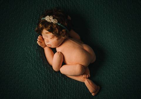 Boneca reborn Jolie em pose lateral clássica deitada sobre manta verde, usada para treino de posicionamento fisiológico em ensaio newborn.'