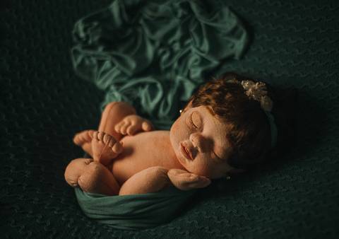 Boneca reborn em pose embrulhada com os pés à mostra, simulando técnica de pose frog adaptada para ensaio newborn com tecido auxiliar.'