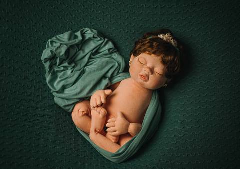 Boneca reborn Jolie deitada com wrap verde e tecido aberto ao lado, posicionada para estudo de composição e iluminação em newborn real.

'