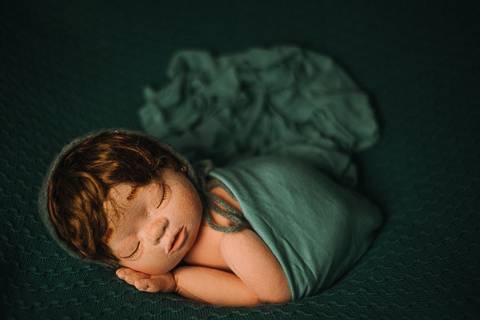 Boneca reborn envolta em manta verde, simulando pose de recém-nascido em ensaio fotográfico profissional com foco em fisiologia e segurança.'