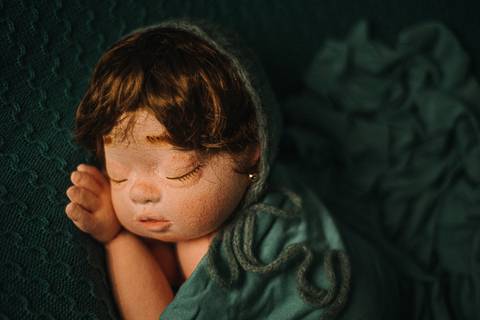 Close da boneca reborn Jolie com gorro de lã e tecido verde, usada para prática de poses fisiológicas e estudo de ângulo na fotografia newborn.'