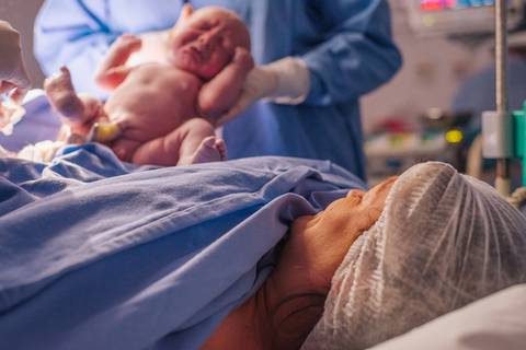 Bebê apresentada à mãe ainda na sala cirúrgica, fotografia de nascimento em cesariana.'