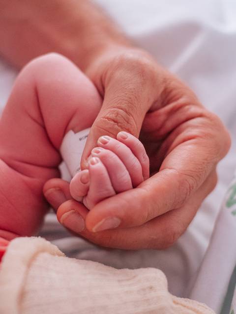 Detalhe das mãos do bebê recém-nascido seguradas pelos pais, fotografia documental de parto.'