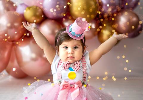 Ensaio Smash the Cake com bebê comemorando 1 ano, braços erguidos, cenário delicado com balões e iluminação suave, fotografia infantil.'