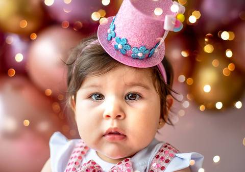 Retrato de bebê em ensaio Smash the Cake, expressão curiosa, cenário clean com balões ao fundo, fotografia de aniversário infantil.'