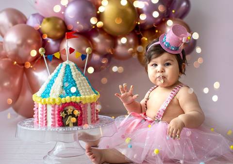 Ensaio Smash the Cake com bebê interagindo com bolo colorido, vestido delicado e cenário com balões.'