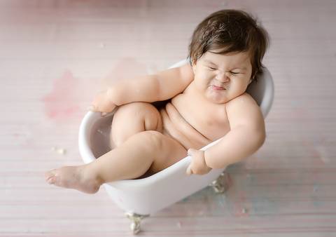 Ensaio Smash the Cake com bebê relaxada em banheira cenográfica, cenário claro e composição delicada para aniversário de 1 ano.'